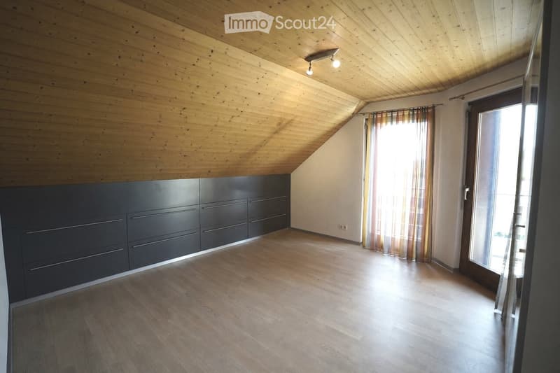 Empty attic room with wooden ceiling and floor, built-in dark cabinets, large window with curtains, and modern lighting fixture.