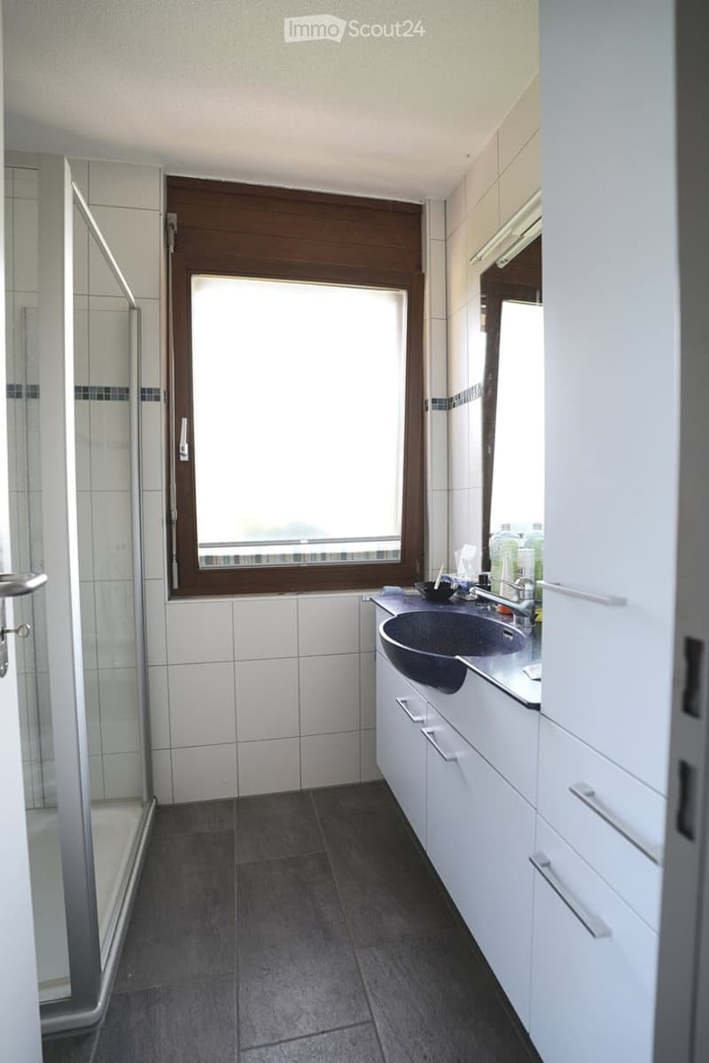 Modern bathroom with white tiles, dark grey floor, white cabinetry, sink, mirror, and a glass-enclosed shower area.