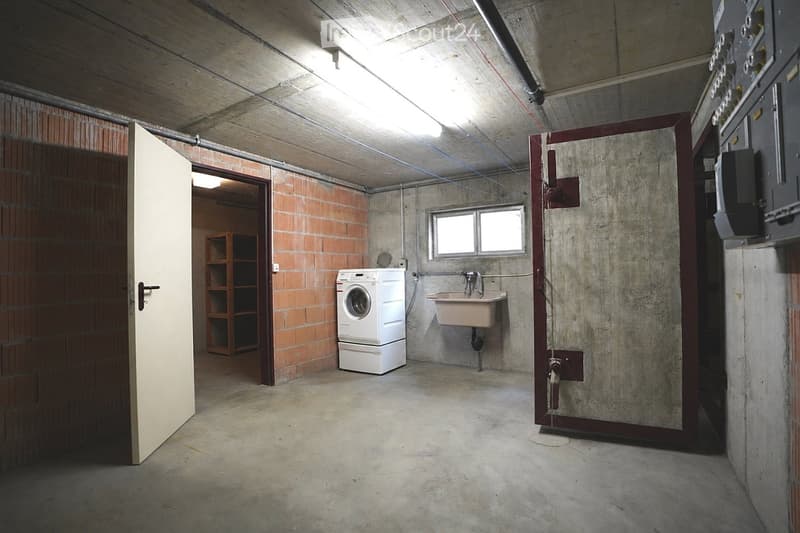 Utility room with a washing machine, sink, and shelves, featuring exposed brick and concrete walls.