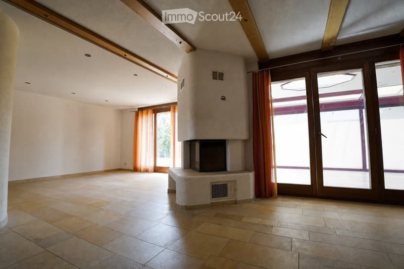 Empty room with tiled floor, exposed wooden beams, large windows with orange curtains, and a built-in fireplace.