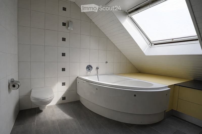 Modern bathroom with a corner bathtub, toilet, and sleek white tiles. Features a skylight window for natural light and a built-in shelf.