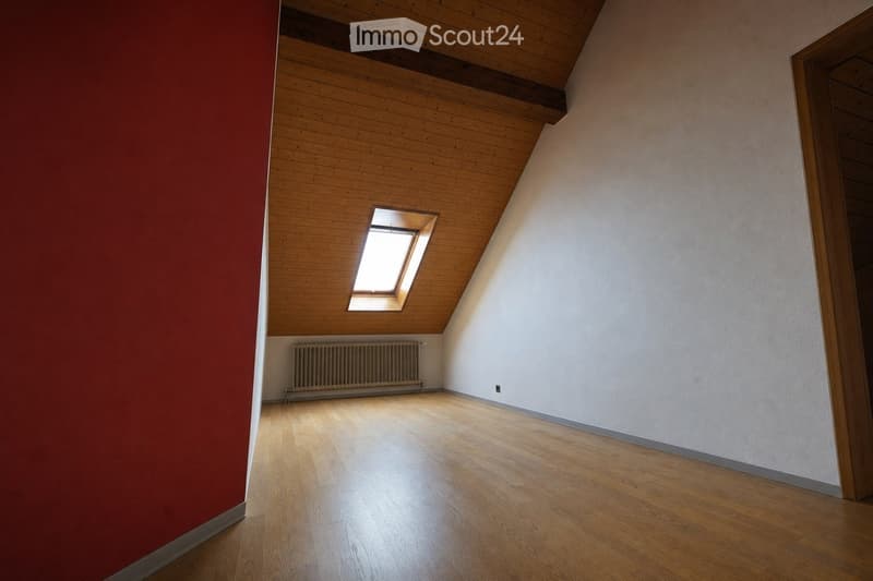 Empty attic room with wooden floor, slanted wooden ceiling, skylight window, radiator, and white walls.