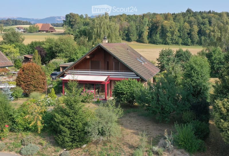 A two-story wooden house with a red balcony and a covered patio area, surrounded by a well-maintained garden with various trees and shrubs, located in a rural area with open fields and distant mountains.