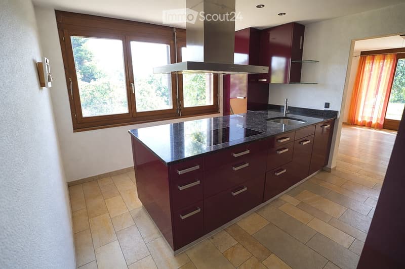 Modern kitchen with dark red cabinets, granite countertop, stainless steel sink, and range hood. Large windows provide natural light.