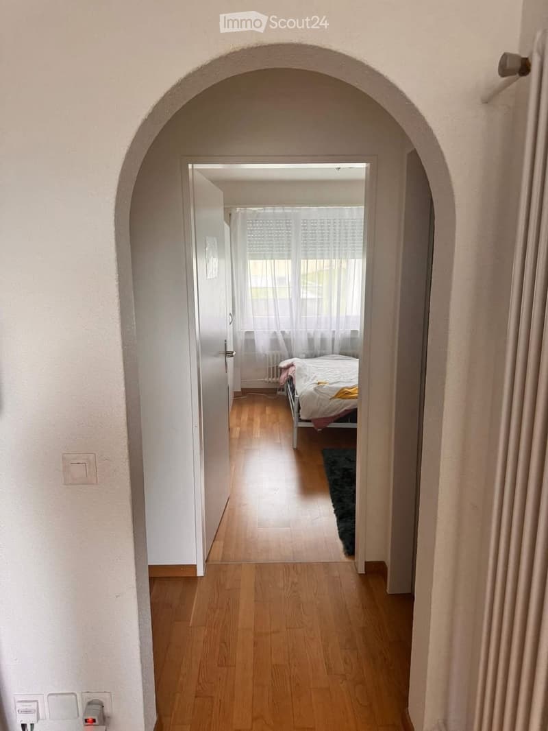 A hallway with wooden flooring and white walls, leading to a bedroom with a bed, rug, and window with curtains.