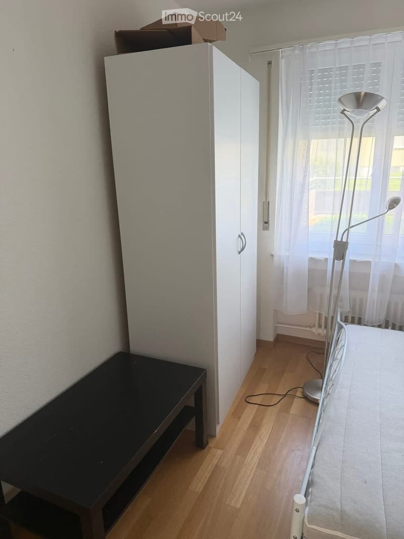 A small, furnished room with a wooden floor, a white wardrobe, a black table, and a floor lamp near the window with sheer curtains.