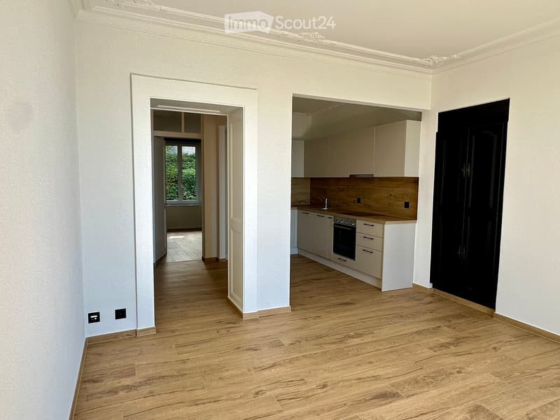 Empty room with light wooden flooring, white walls, and a doorway leading to another room with a window and a kitchen area visible in the background.