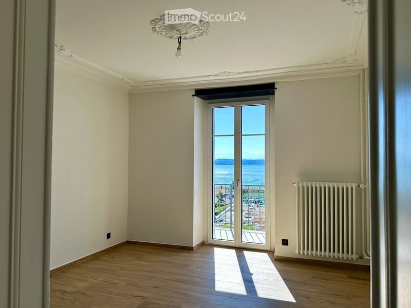 Empty room with wooden flooring, white walls, large sliding glass doors leading to a balcony with a sea view, and a radiator on one wall.