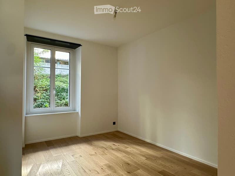 Empty room with light-colored wooden flooring and white walls, featuring a large window with a view of greenery outside.