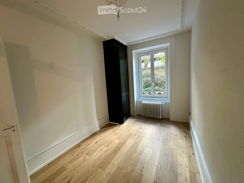 Empty room with light wooden flooring, white walls, a window with a radiator below, and a black wardrobe. No furniture or personal items are present.