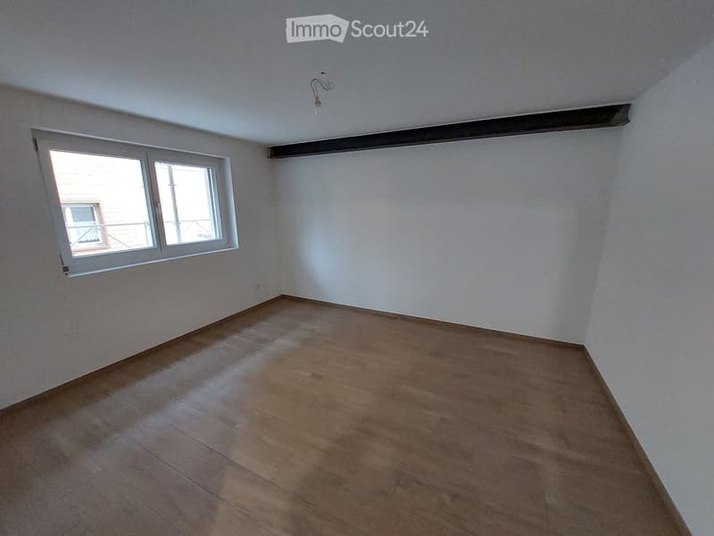 Empty room with wooden flooring and white walls, featuring a single window with white frame and a visible electrical outlet on the wall.