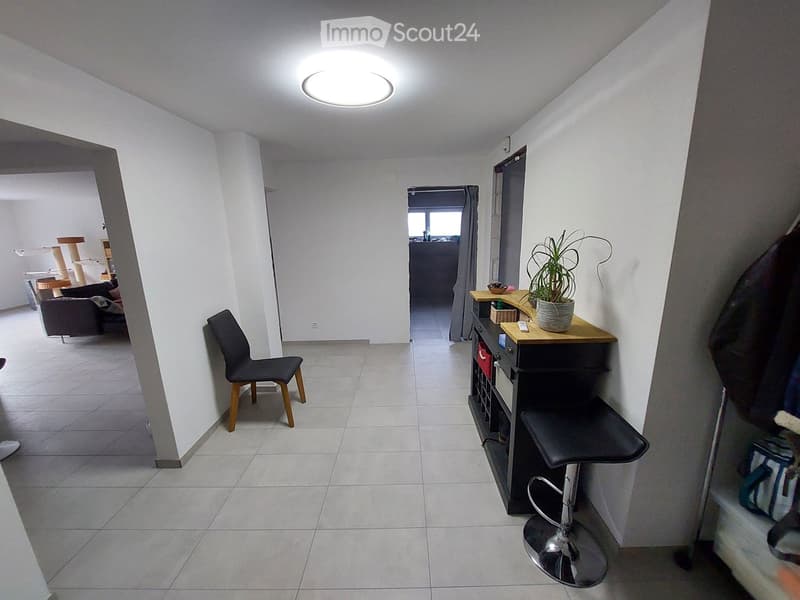 A modern, well-lit hallway with tiled flooring, a round ceiling light, and a small seating area with a chair and a stool. A small table with a potted plant is visible, and a cat tree and sofa are in the adjacent room.