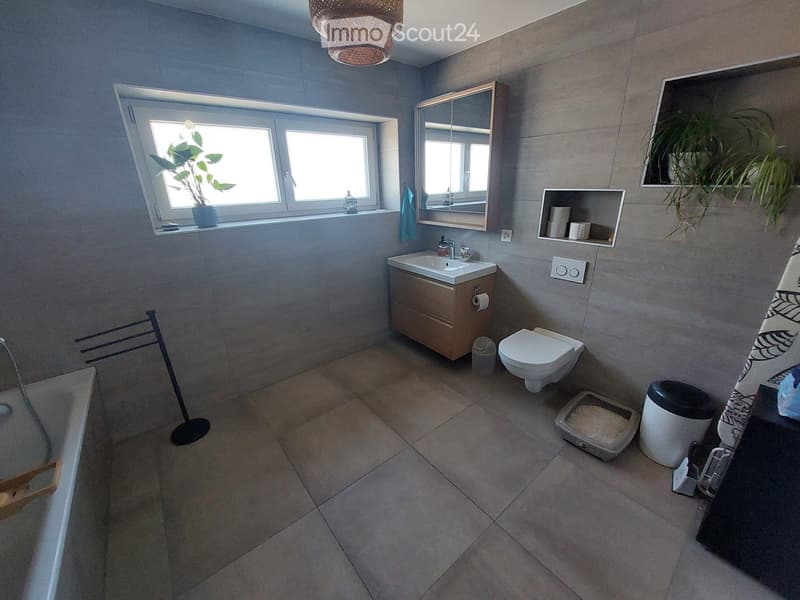 Modern bathroom with grey tiled walls and floor, featuring a sink with a wooden cabinet, a toilet, a bath, a mirror, a towel rack, a cat litter box, and a plant on the windowsill.