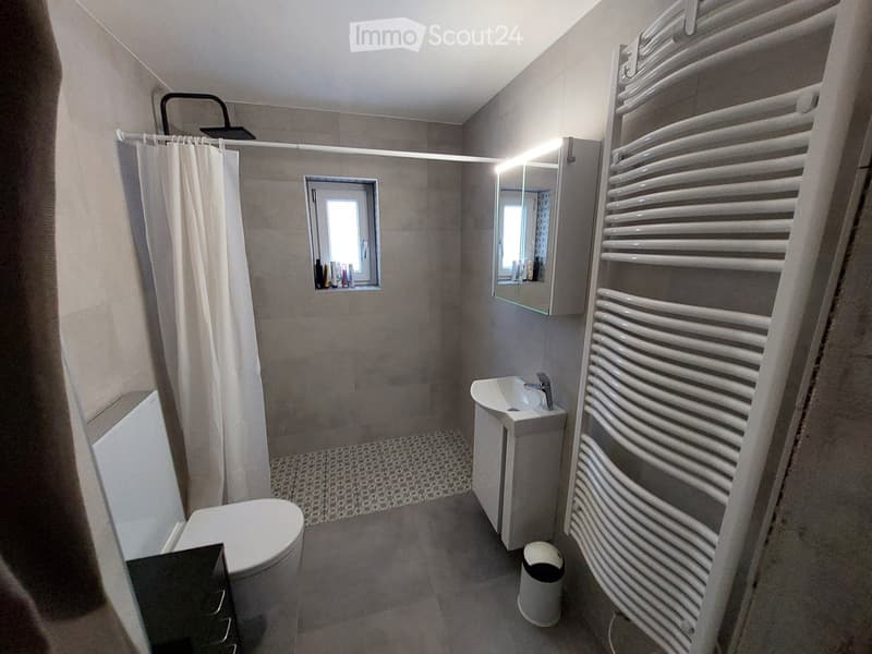 Modern bathroom with a toilet, sink, mirror, radiator, and shower curtain. Features a small window and a built-in shelf with toiletries.