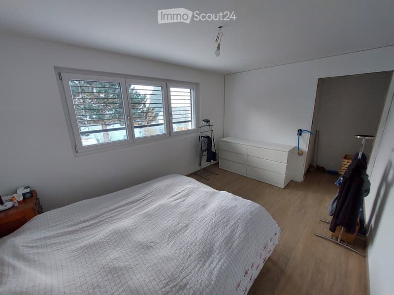 Bright bedroom with wooden flooring, large window with blinds, bed with patterned bedding, nightstand, drawer unit, and coat rack.