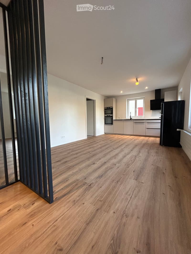 Open-plan living and dining area with light wooden flooring, modern kitchen area featuring white cabinets, stainless steel appliances, and a large window.