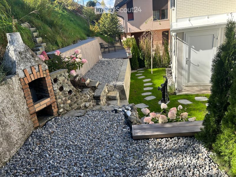 Garden with gravel pathway, stone steps, small wooden bench, stone fireplace, potted plants, and a small grassy area with stepping stones.