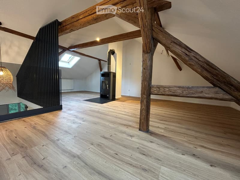 Empty attic space with wooden beams, wooden flooring, and a modern black wood-burning stove. Features exposed wooden beams, a skylight, and a modern staircase with a glass railing.