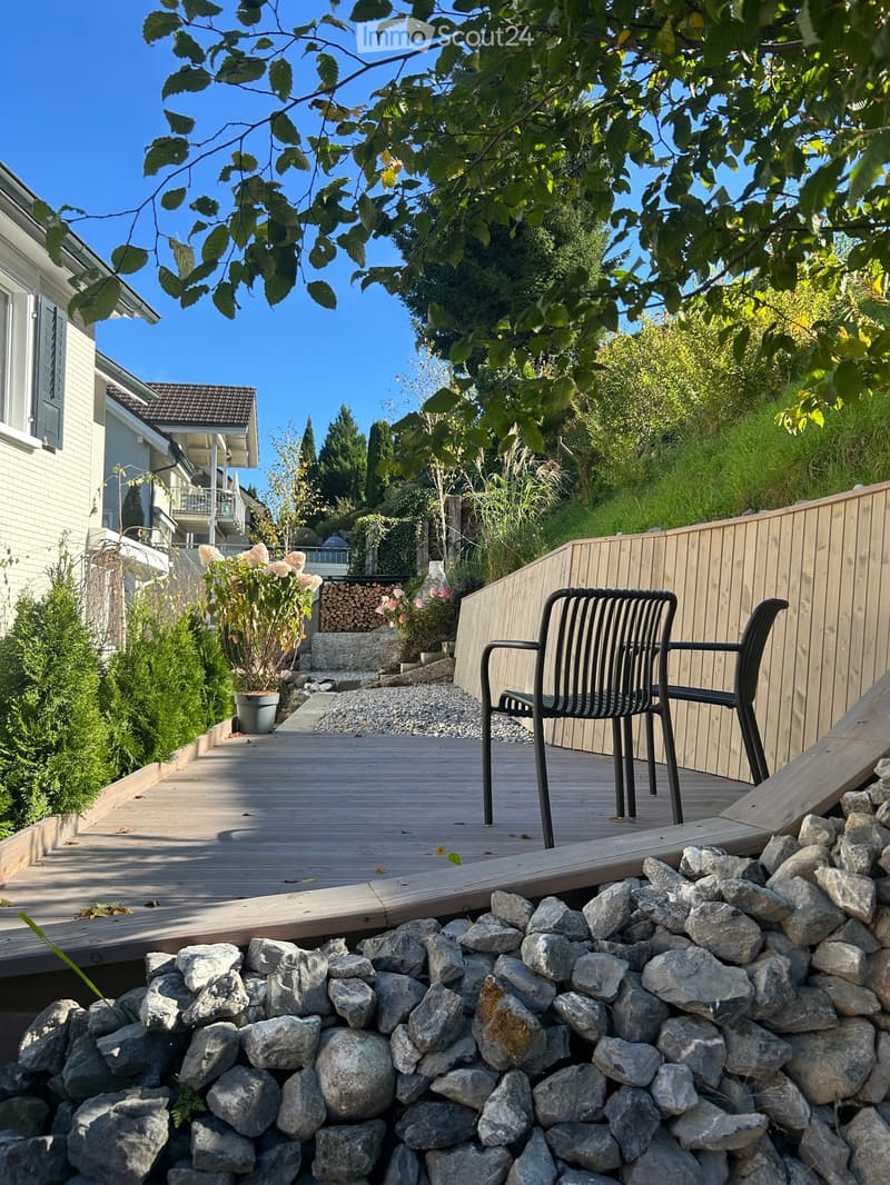 Wooden deck with a black metal chair, surrounded by a stone border and lush greenery, with a house and trees in the background.
