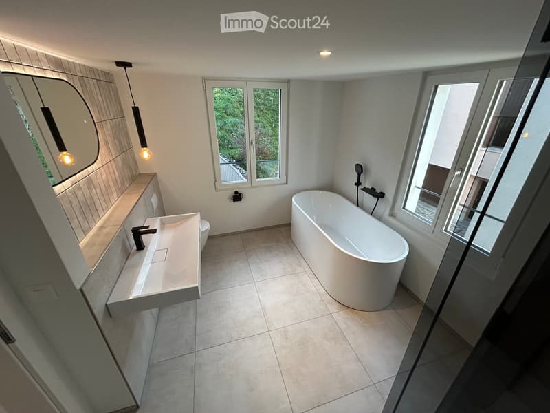 Modern bathroom with a freestanding bathtub, a sink with a sleek faucet, a large mirror, and a toilet. The space features large windows and pendant lights.