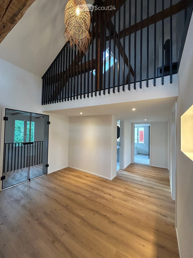 Spacious hallway with wooden flooring, modern staircase with black railings, and a hanging woven light fixture. The hallway features white walls and recessed lighting, with a glass door leading to an outdoor area.