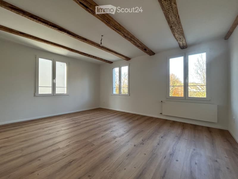 Empty room with wooden flooring, white walls, exposed wooden beams on the ceiling, and multiple windows allowing natural light.