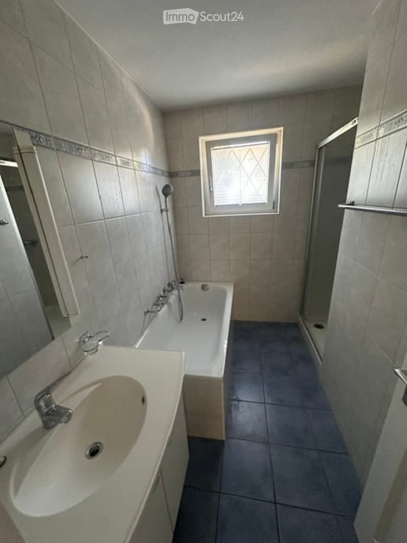 Bathroom with a white sink, bathtub, mirror, and a small window above the bathtub. The walls are tiled, and the floor is dark blue tiles.