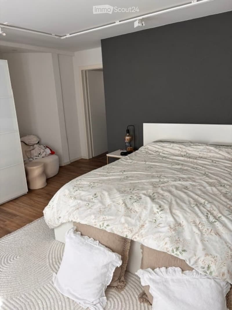 Modern bedroom with a large bed, white headboard, nightstand with a lamp, and a wardrobe. The room features wooden flooring and a dark accent wall.