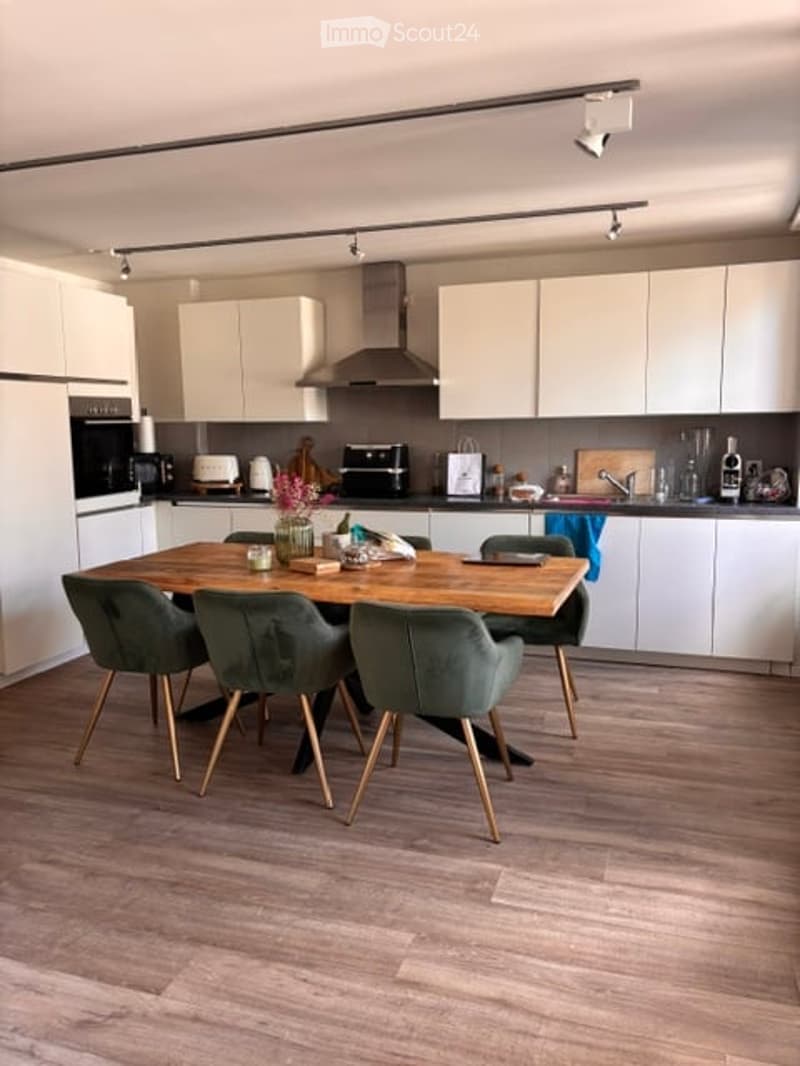 Modern kitchen with white cabinets, wooden dining table, four green chairs, and various appliances including an oven and toaster. The floor is light wood, and the space is well-lit with track lighting.