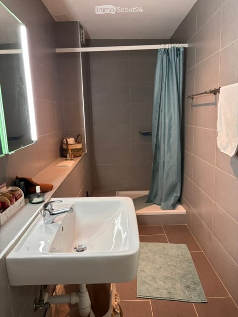 Modern bathroom with a white sink, chrome faucet, shower with a blue curtain, and a towel on a rack. A small basket and a rug are also visible.