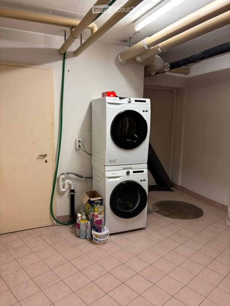 Utility room with stacked washing machine and dryer, cleaning supplies on floor, tiled floor, exposed pipes, and a door.