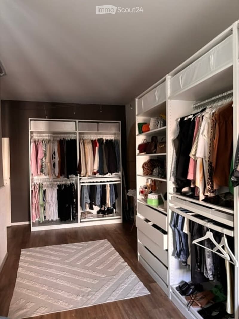 Spacious walk-in closet with built-in wardrobes, clothing racks, and drawers. The floor is wooden, and a large rug is placed in the center.