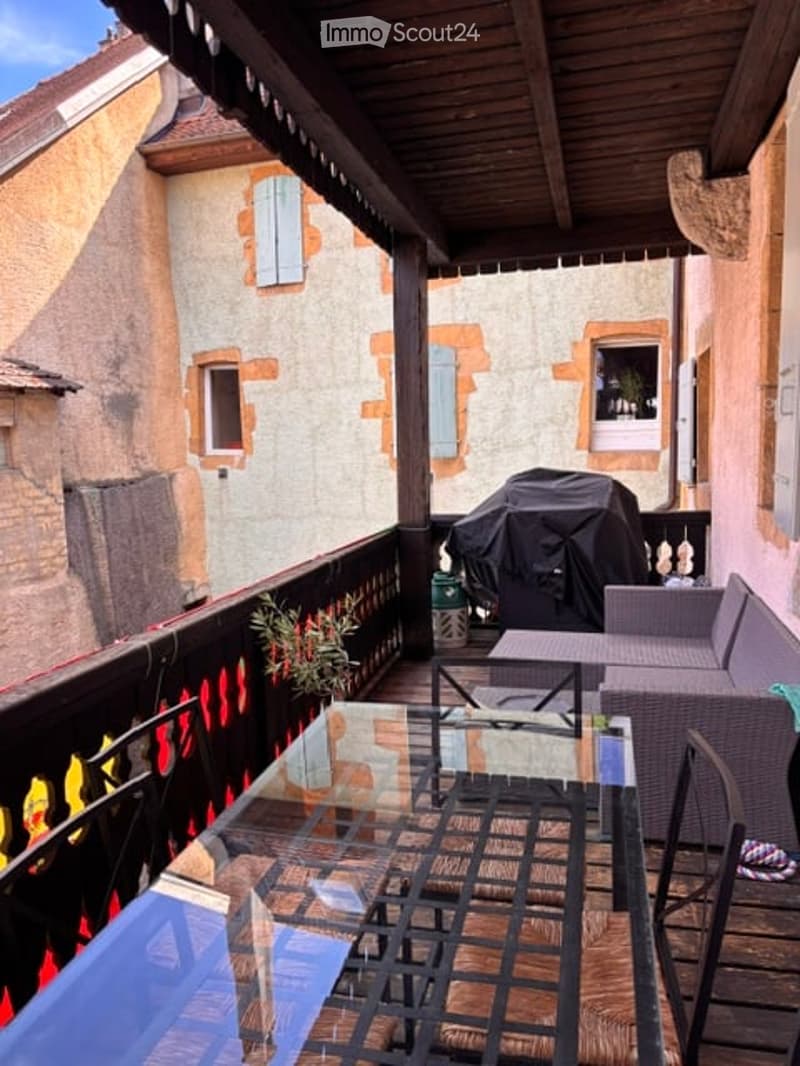 Balcony with wooden decking, glass table, chairs, sofa, and covered grill, overlooking a historic building with stone walls and shutters.