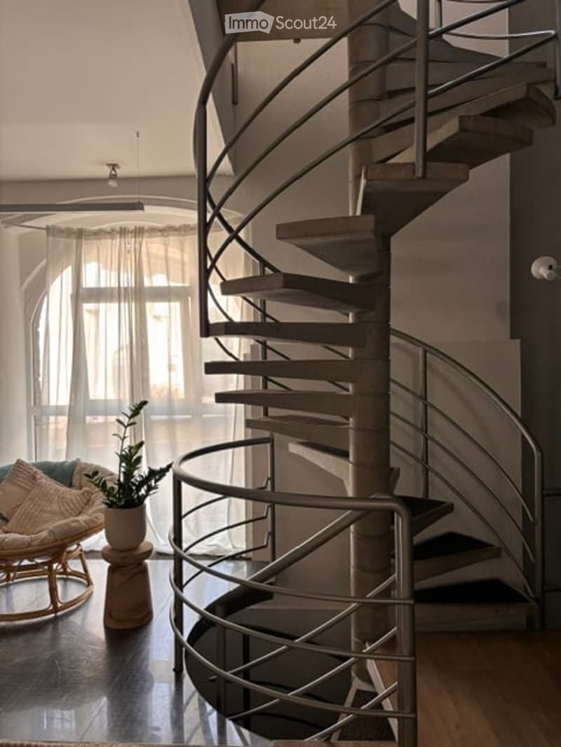 Modern interior with a spiral staircase, a small seating area featuring a wicker chair and a potted plant on a stool, and large windows with sheer curtains.