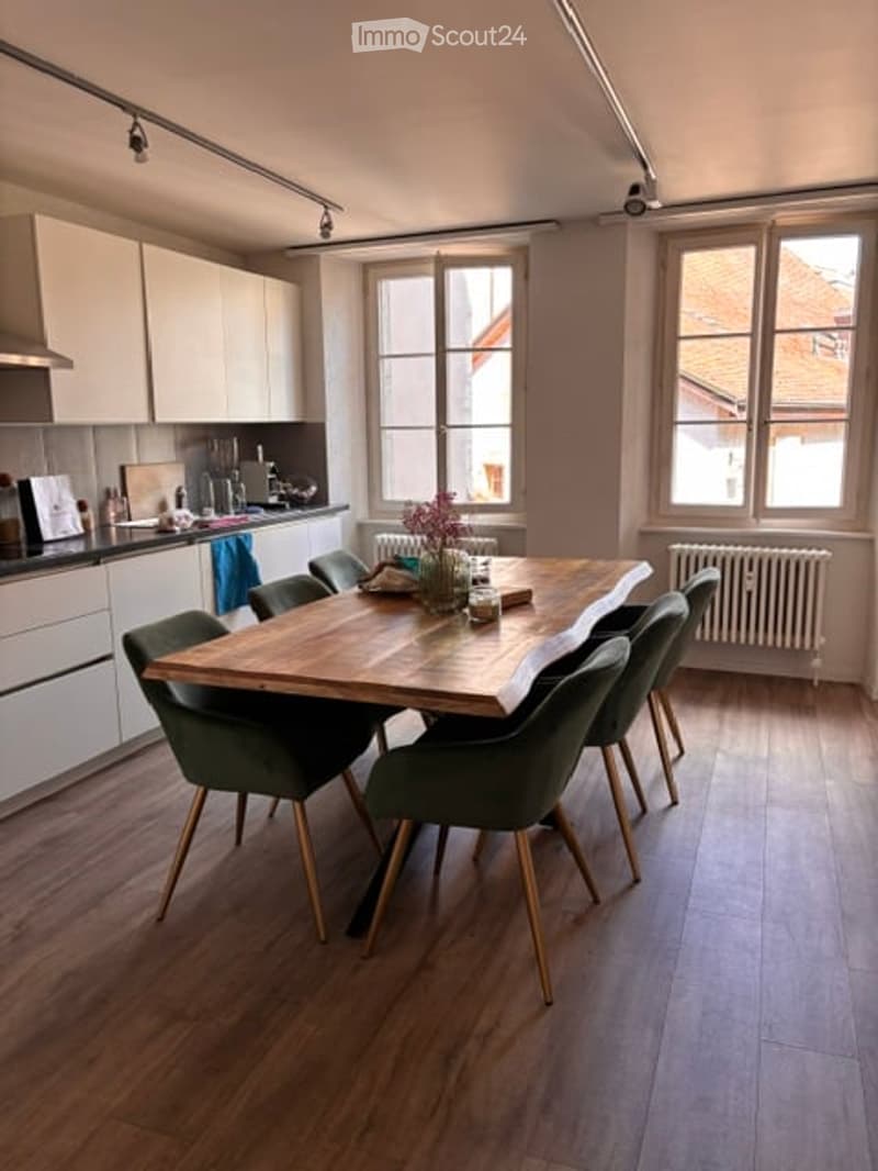 Bright, modern dining area with a large wooden dining table surrounded by green chairs, white kitchen cabinets, and a sink area. Natural light streams in through large windows, and a vase with flowers adds a touch of color.