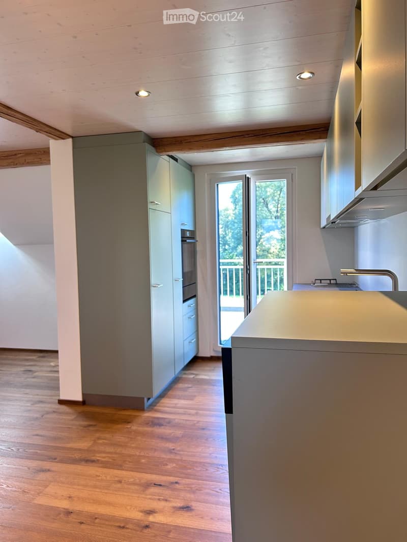 Modern kitchen with light grey cabinets, wooden flooring, and a view of the outdoor area through large glass doors.
