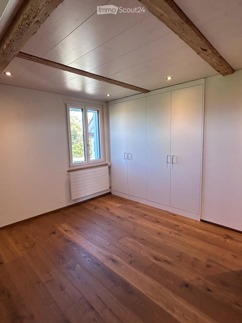Empty attic room with wooden flooring, white walls, built-in wardrobes, and a window with a radiator below it.