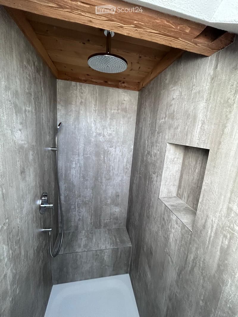 Modern bathroom with grey stone walls, a sleek showerhead mounted on a wooden shelf, and a built-in niche for storage.