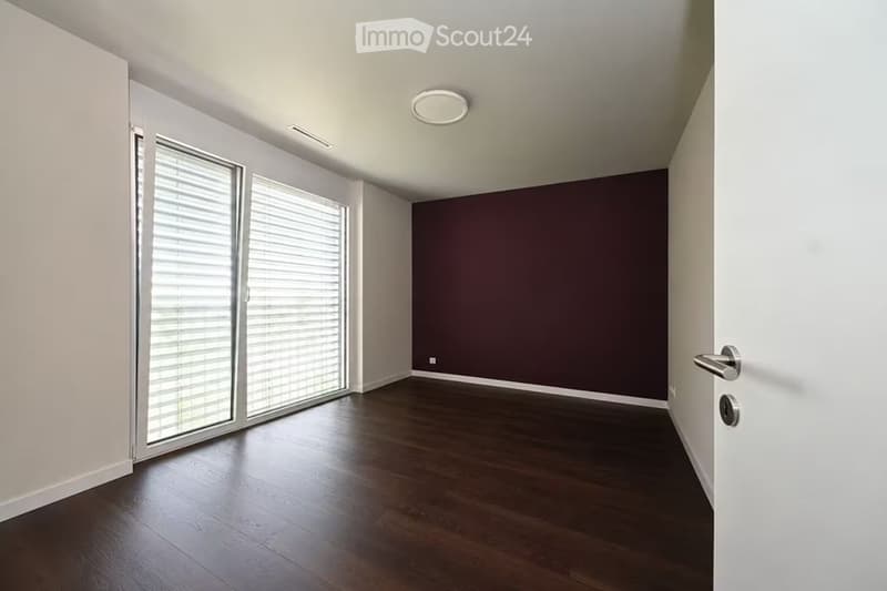 Empty room with dark wooden flooring, white walls, large window with blinds, and a door.