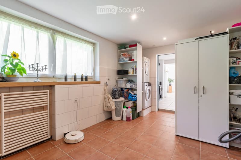 Well-lit laundry room with tiled walls and floor, featuring a washing machine, cabinets, a radiator, a candle holder, a plant on the windowsill, and a vacuum cleaner on the floor.