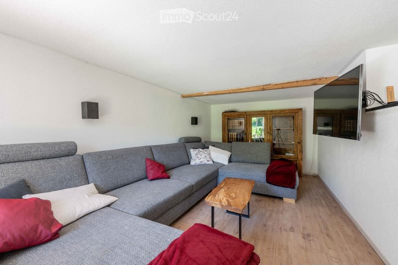 Modern living room with a grey sectional sofa, wooden coffee table, wooden cabinet, and wall-mounted television. The room features light wooden flooring and white walls.
