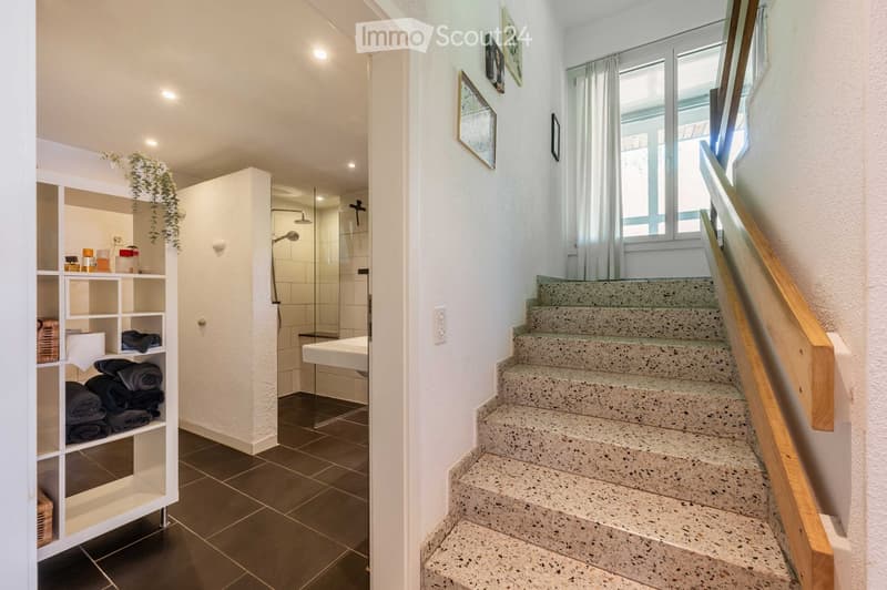 Modern hallway with a staircase leading upwards, featuring a wooden handrail and a glass shower enclosure visible in the background. The hallway has white walls, a dark tiled floor, and a shelf with towels and decorative items.