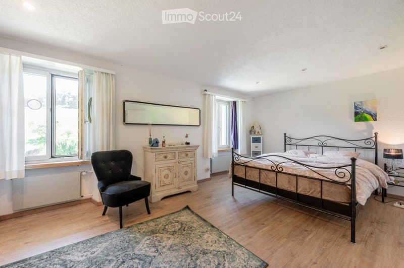 Cozy bedroom with wooden flooring, large bed with metal headboard, black armchair, dresser with mirror, and a small table lamp on a nightstand.