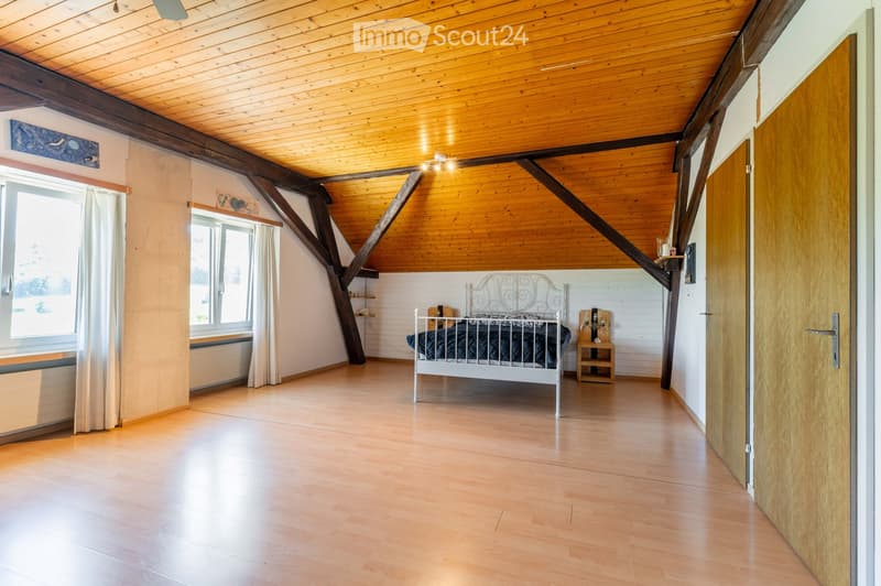 Bright bedroom with wooden floors, slanted wooden ceiling, large windows with white curtains, a white metal bed, a small wooden nightstand, and a shelf with decorative items.