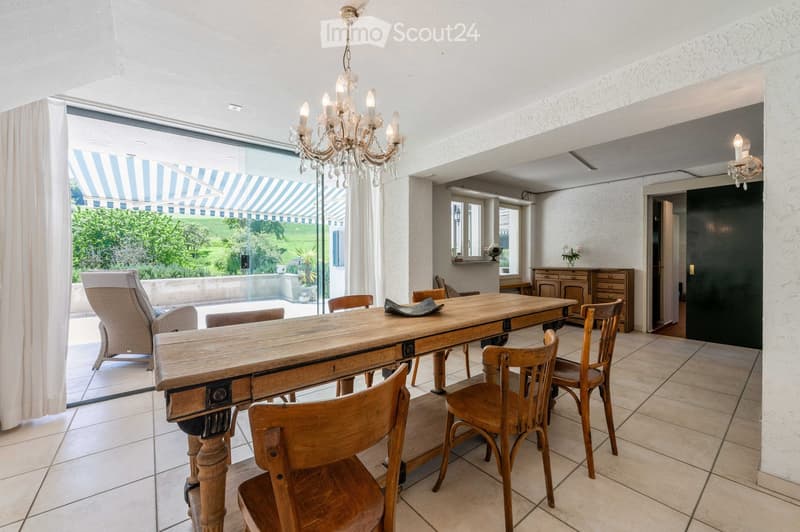 Bright dining area with a long wooden dining table surrounded by wooden chairs, a wicker sofa, and a chandelier hanging from the ceiling. Large windows offer a view of a garden and outdoor seating area.