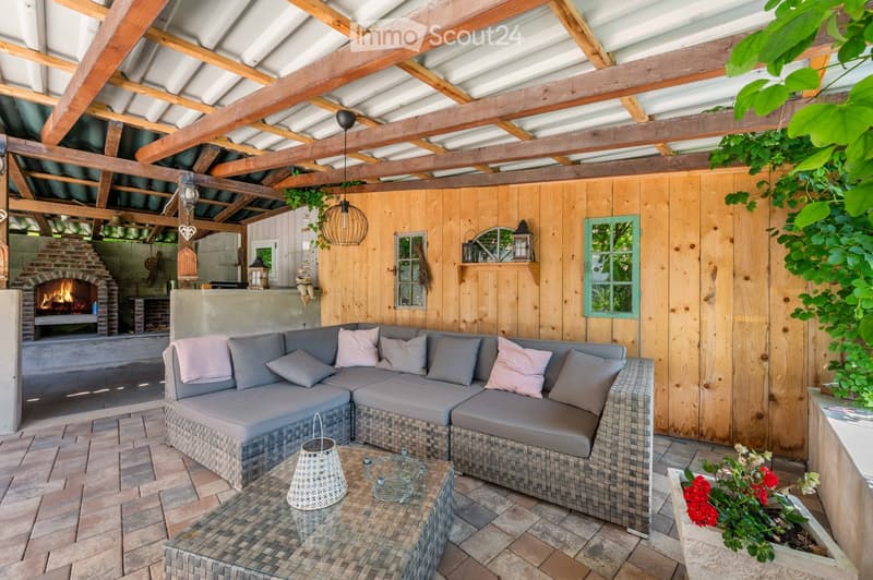 Outdoor patio with wooden walls, exposed wooden beams, grey wicker sofa, coffee table, and a brick fireplace.