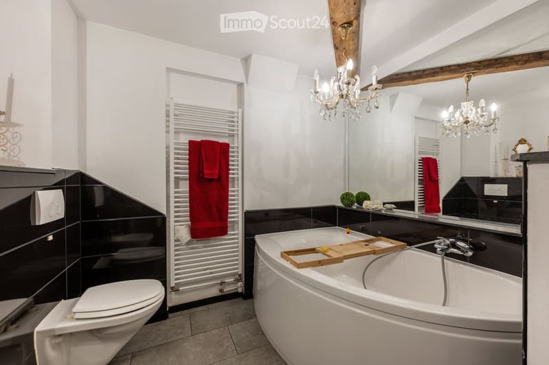 Modern bathroom with a freestanding bathtub, toilet, towel rack with red towels, large mirror, chandelier, and decorative items on the counter.