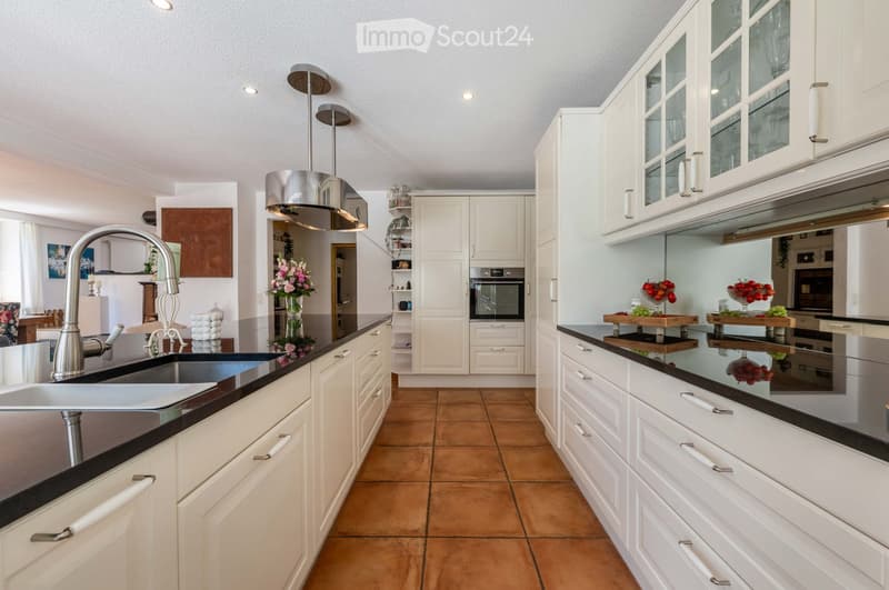 Modern kitchen with white cabinets, stainless steel appliances, and a central island. Features include a large sink with a modern faucet, built-in oven, and decorative fruit bowls on the island.
