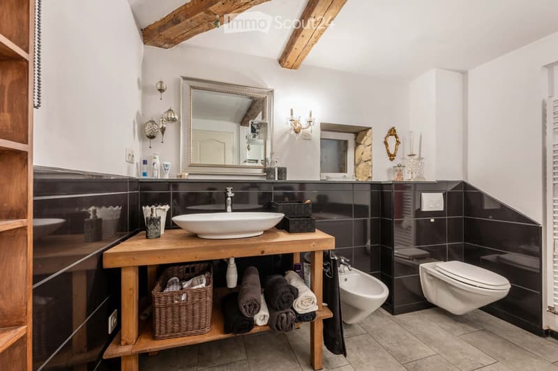 Modern bathroom with a wooden vanity, white sink, toilet, bidet, and decorative mirrors. Features exposed wooden beams, dark tiled walls, and neatly arranged towels and candles.