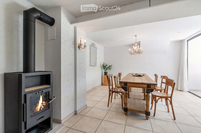 A cozy dining area with a wooden dining table and chairs, a black wood-burning stove with a visible flame, a chandelier, and a small plant on a side table.